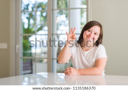 Down syndrome woman at home showing and pointing up with fingers number three while smiling confident and happy.