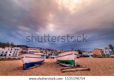 View on small shingle beach with two boats in front and small town in distance under stormy sky before sunset in Costa Brava (Spain)