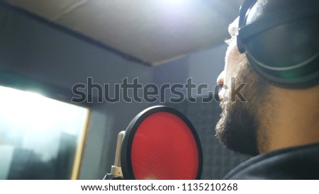 Male singer with beard in headphones singing song into the microphone at sound studio. Young man recording new melody or song. Working of creative musician. Show business concept. Slow motion.
