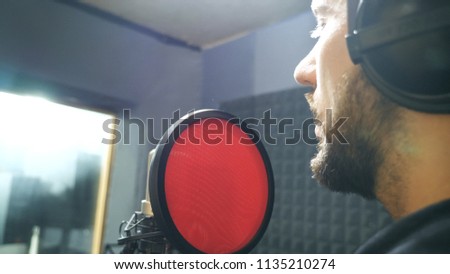 Male singer with beard in headphones singing song into the microphone at sound studio. Young man recording new melody or song. Working of creative musician. Show business concept. Slow motion.
