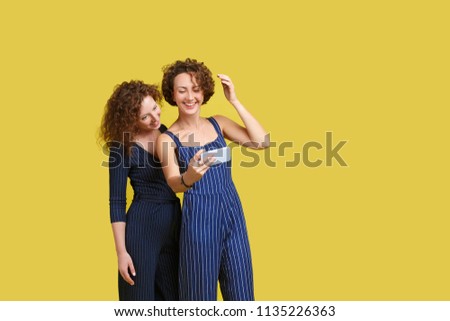 Pleased caucasian ginger girl gently smiling while her European friend making selfie. Indoor portrait of glad sisters with smartphone taking picture of herself on yellow background. Copy space
