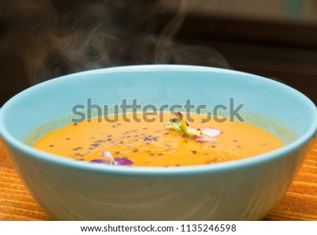 One bowl with steaming hot pumpkin soup in a light blue bowl garnished with fresh pansy flowers and black sesame seeds - Healthy eating and living concept background. Autumn season vegetables cooking.