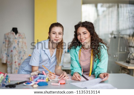 Young pretty tailors happily looking in camera drawing sketches of new clothes together in cozy sewing workshop
