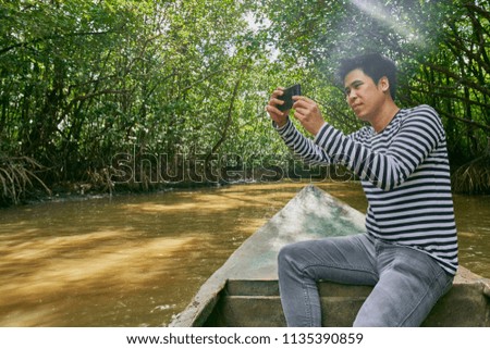 Happy black and white shirt Male Traveler uses camera for taking the beautiful sky and natural view photo while sailing on the boat along the river to visit nature Mangrove Ecosystem environment