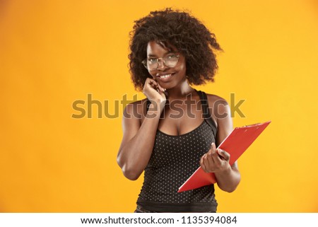 The portrait of a beautiful friendly African American woman with a curly afro hairstyle and red folder isolated on a orange studio background