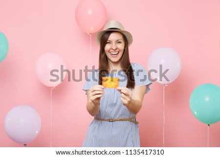 Portrait of laughing fascinating young woman in straw summer hat and blue dress holding credit card on pastel pink background with colorful air balloons. Birthday holiday party people sincere emotion