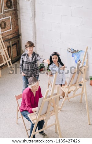 Full length portrait of  female art teacher watching group of students painting at easels in art class, copy space