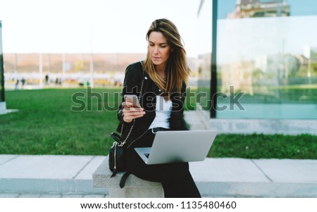 Beautiful caucasian female waiting for sms on her mobile phone to confirm transaction for online payment for the flight tickets. Student girl reading emails on a smartphone while sitting with a laptop Royalty-Free Stock Photo #1135480460