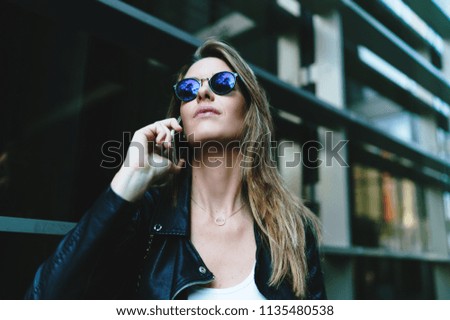 Closeup photo of young beautiful caucasian female in sunglasses wearing black leather jacket and white t-shirt talking by a mobile phone while standing on a blurred business center background.