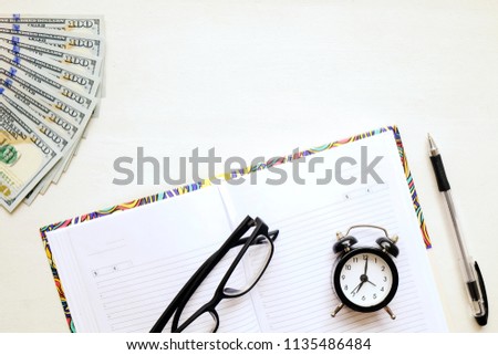 Time and money management concept. Open blank notebook, alarm clock, pen, hipster eyeglasses with black frame, multiple one hundred dollar bills pack. 