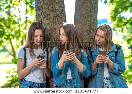 Three girls schoolgirl teen. In summer park by tree. He holds smartphone in his hands, writes messages, reads correspondence. The concept of children in social networks, communication on Internet.