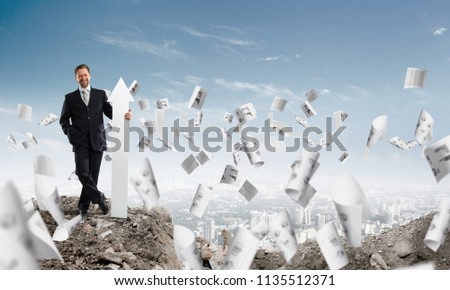 Horizontal shot of confident business man holding white arrow while standing among flying papers in ruins with cityscape view on background.
