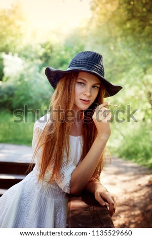 Portrait of a red-haired girl with long hair, in a black wide-brimmed hat. Bites his teeth a blade of grass. Warm sunlight