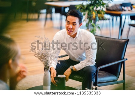 A handsome and confident Chinese Asian business man sits on the chair and has a chat with his team during the day. He is enjoying a hot beverage while he talks animatedly to his colleagues.