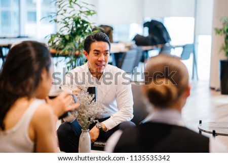 A handsome and confident Chinese Asian business man sits on the chair and has a chat with his team during the day. He is enjoying a hot beverage while he talks animatedly to his colleagues.