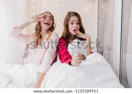Lovely curly girl in eyemask playfully posing while her friend making selfie. Good-looking female models lying in bed and taking picture of themselves.
