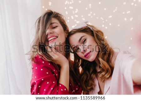 Close-up portrait of winsome curly woman making selfie with friend. Indoor photo of carefree girls taking pictures of themselves.