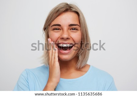 Cheerful beautiful young woman with blonde hair and braces on teeth laughing and touching her face by hand isolated over white background Royalty-Free Stock Photo #1135635686