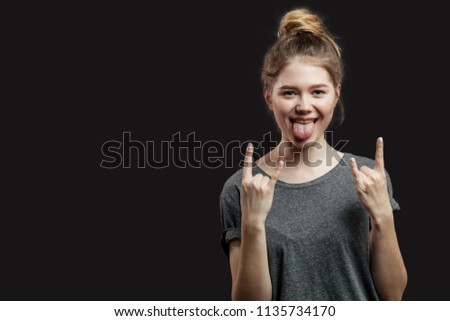 Isolated studio shot of young hipster female looking at camera and making rock sign with both hands, keeping her tongue out of mouth while standing against black background