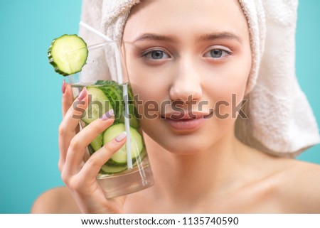 Beautiful caucasian woman with healthy young skin having towel on her head pressing a glass of cucumber water to her face as if refreshing herself from the heat of the day. Relaxation SPA concept. Royalty-Free Stock Photo #1135740590