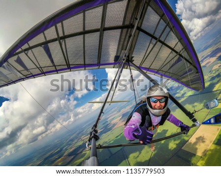 Smiling girl hang glider pilot shows thumb up while flying high over green fields below clouds. Wide angle selfie photo of extreme sport taken with action camera Royalty-Free Stock Photo #1135779503