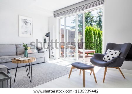 Armchair with patterned pillow and stool in living room interior with grey sofa and posters. Real photo