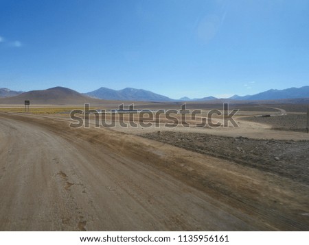 Picture from a touristic small bus of a road covered by sand, the blue sky, and the Andes mountains driving through the Atacama desert in Chile towards the Bolivian high plateau in South America.