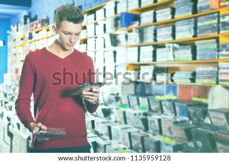 Handsome focused young guy looking on box of DVD movie during shopping in CD shop