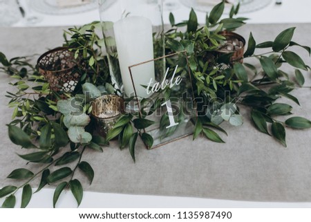 on table with gray tablecloth is a candle in candlestick, plate with number and composition of greenery Royalty-Free Stock Photo #1135987490