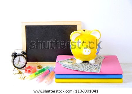Savings for education concept. Yellow piggy bank standing on stack of colorful notebooks & pack of new crispy one hundred dollar bills. School supplies, blank blackboard, clock. Background, copy space