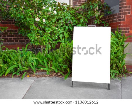 Blank white outdoor advertising stand mock up template. Clear street signage board placed against green plants growing at front of old brick house. 