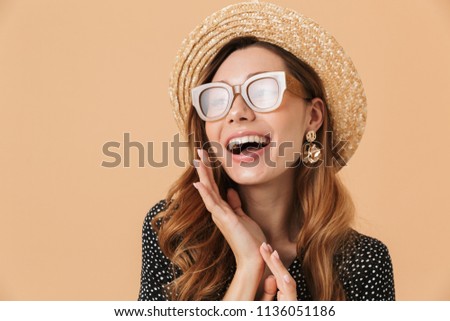 Portrait of gorgeous stylish woman wearing straw hat and sunglasses smiling and looking aside isolated over beige background