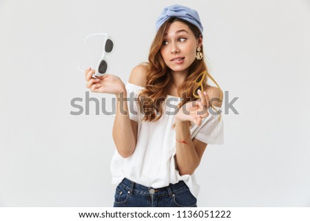 Portrait of stylish indecisive woman 20s doubting while holding two pairs of sunglasses isolated over white background