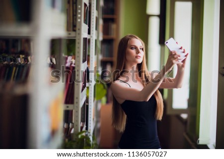 Image young smiling woman student sitting in library make selfie with peace gesture by mobile phone. Looking aside.