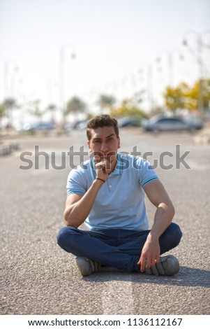 Outdoor shot of young man sitting crossed legs on the asphalt in the park puts hand on chin thinking, blurred view behind him of stopped cars on the background.