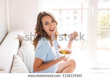 Cheerful young woman eating healthy breakfast while sitting on a couch at home Royalty-Free Stock Photo #1136119265