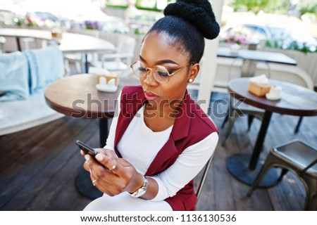 African american girl wear in glasses with mobile phone sitting at outdoor caffe.