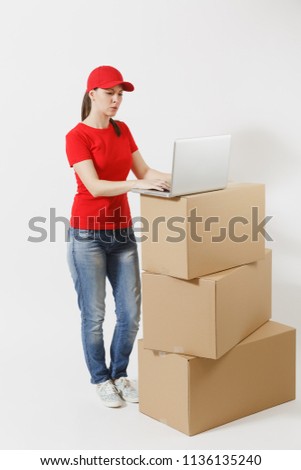 Full length portrait of delivery woman in red cap, t-shirt isolated on white background. Female courier standing near empty cardboard boxes with laptop pc computer. Receiving package. Copy space