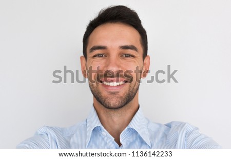 Closeup portrait of happy handsome young male smiling with healhty toothy smile looking at the camera, making self portrait. Cheerful unshaven man wearing blue shirt posing in studio background.People