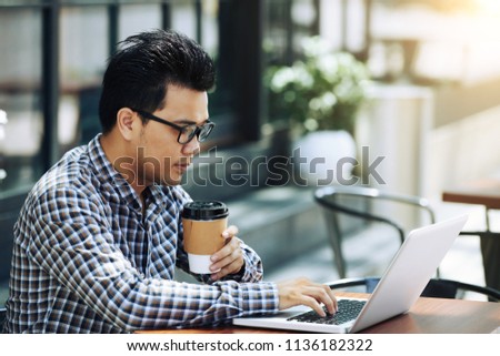 Asian young man drinking take-out coffee and working on laptop