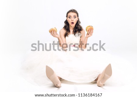 shocked young bride in wedding dress sitting on floor with burgers in hands on white