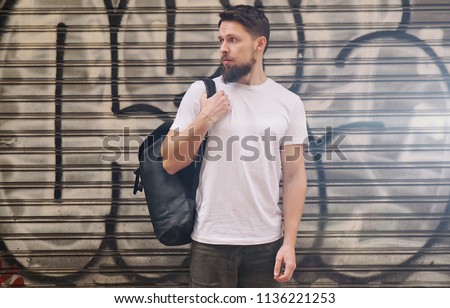 Young man with a black backpack in a white t-shirt standing near the graffiti wall. Horizontal mock-up.