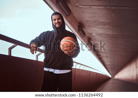 Dark-skinned bearded guy dressed in a black hoodie and sports shorts leaning on a guardrail while standing with basketball.