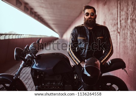 Stylish fashionable biker dressed in a black leather jacket with sunglasses holds a helmet sitting on his custom-made retro motorcycle, looking at camera.