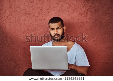 Close-up portrait of an African-American bearded guy dressed in a white shirt and sports shorts holds on a laptop while leaning on a wall, looking at camera.