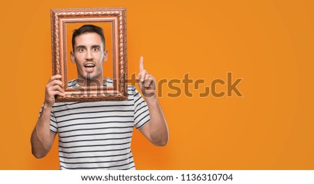 Handsome young man looking through vintage art frame surprised with an idea or question pointing finger with happy face, number one