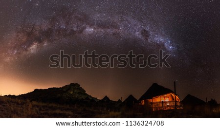 A panorama of the purple magic milky way and a sky full of stars above a little African hut which is shining in orange light with a little hill in the background and more houses in the back