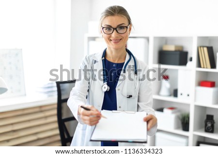 A young girl in a white coat is standing near a table in her office and holding a tablet and a pen. A stethoscope hangs around her neck.