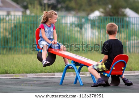 Two young blond children preschooler girl with long ponytail and cute school boy swing on see- saw on bright green blurred background. Joys and games of childhood, outdoors activities concept.