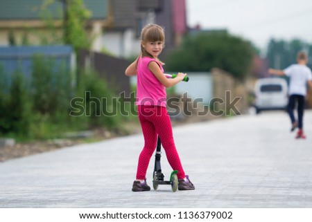 Pretty young long-haired blond child girl in pink clothing with scooter on sunny suburb street and boy on skateboard blurred summer bright background. Children activities, games and fun concept.